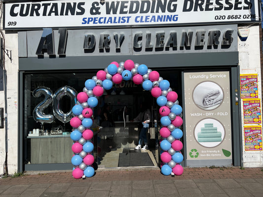 A storefront entrance for a dry cleaning business decorated with a pink, blue, and silver balloon arch. The sign reads 'ALL DRY CLEANERS' and 'Curtains & Wedding Dresses Specialist Cleaning'.
